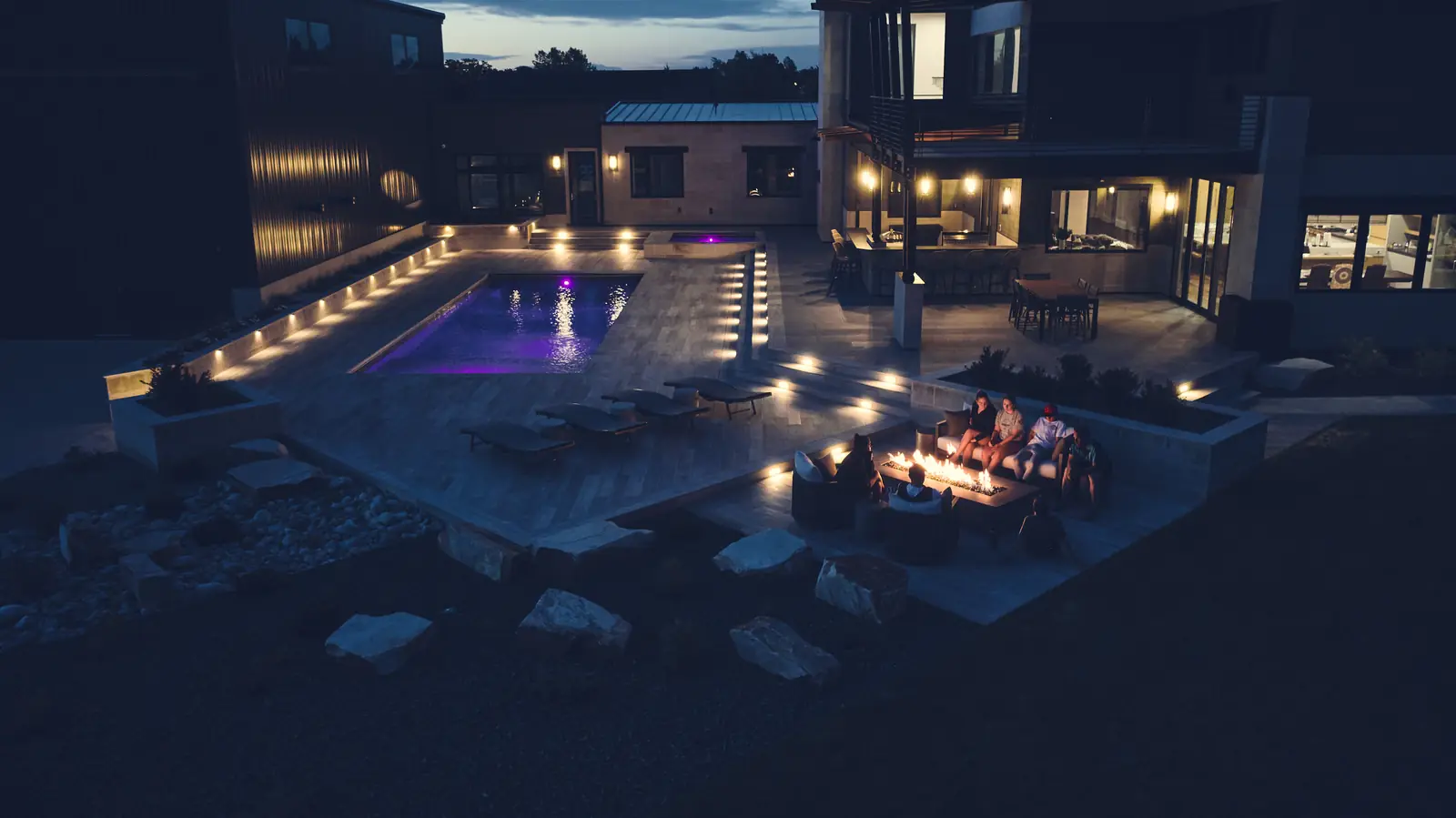 Wide dusk view of the pool, modern home, and fire pit lounge under a fading sky at the Johnson Estate