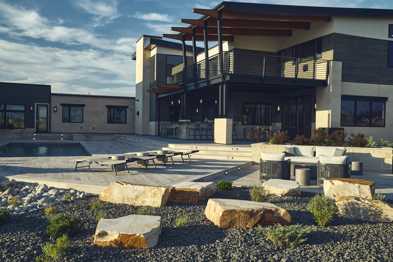 Wide backyard view of a two-story modern home with layered patios, boulder garden, and lounge seating, Colorado Front Range